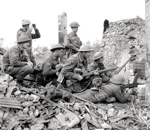 En Italie, un soldat canadien se sert de jumelles pour balayer le champ de bataille alors que d’autres attendent derrière une partie de mur, en décembre 1943. [PHOTO : FREDERICK G. WHITCOMBE, BIBLIOTHÈQUE ET ARCHIVES CANADA — PA136332]