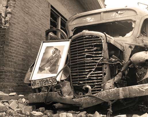 A smashed portrait of Hitler leans against a destroyed German vehicle, May 1945. [PHOTO: LIBRARY AND ARCHIVES CANADA—PA130978]