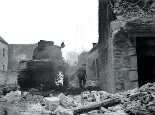 A tank burns while a Canadian picks his way through St. Lambert-sur-Dives. [PHOTO: DONALD I. GRANT, LIBRARY AND ARCHIVES CANADA—PA132192]