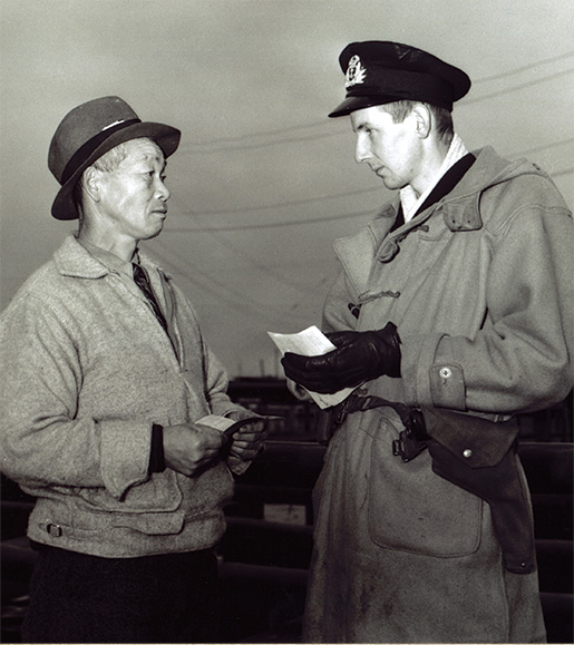 An officer with the Royal Canadian Naval Volunteer Reserve examines the papers of a Japanese-Canadian fisherman in Steveston, B.C., in December 1941. [DND/LAC/PA-170503]